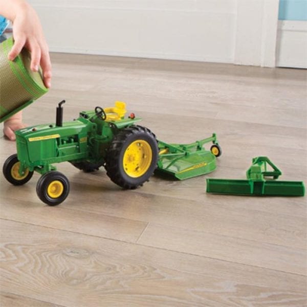Image of the 1/16 scale John Deere Big Farm 4020 toy tractor with rotary cutter and rear blade attachments on the floor by a child.