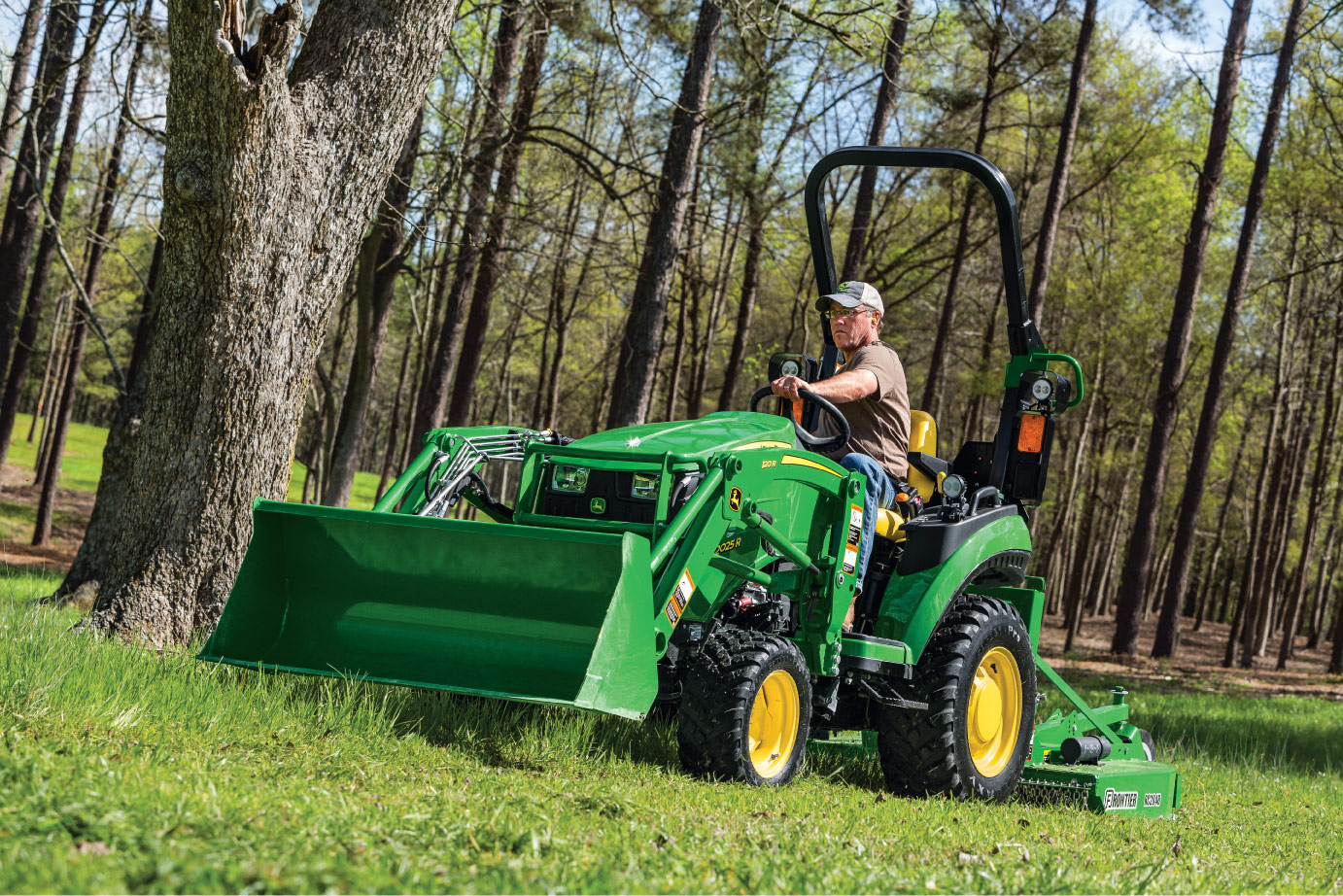 John Deere 2025R compact utility tractor with 120R loader and rotary cutter mowing grass on a rural property.